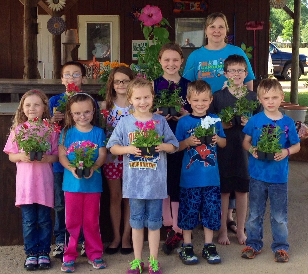 Nine children and one woman with blue shirt standing in front of a brown counter. Children are each holding a four-pack of petunias. Large pink hibiscus flower is behind the group.